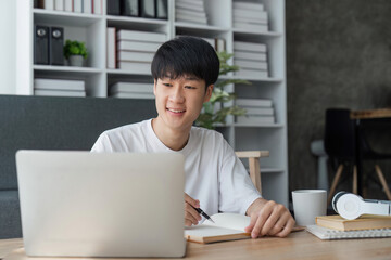 young man studying at home using his laptop, receiving lectures online and taking notes, smiling