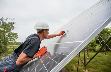 Man engineer installing solar modules. Male worker in safety helmet and work gloves checking middle clamp while mounting solar photovoltaic panel system.