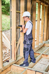 Carpenter constructing wooden framed house. Man worker in glasses working with screwdriver, wearing work overalls and helmet. Concept of modern eco-friendly construction.