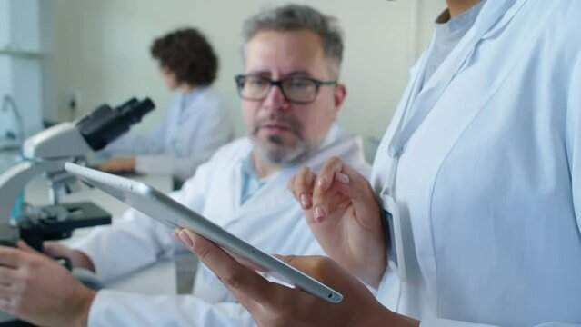 Tilt focus on Biracial woman in medical gown holding tablet and listening to male Caucasian colleague sitting at desk looking at probe through microscope