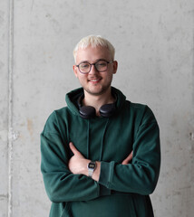 A man with blue hair, eyeglasses, and a green sweatshirt confidently poses with his arms crossed...