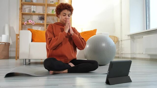 Yoga mindfulness meditation. Young healthy african girl practicing yoga at home. Woman sitting in lotus pose on yoga mat eyes close hands in prayer meditating indoor. Girl doing breathing practice