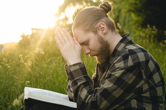 Man praying on the holy Bible in a field during sunset, male sitting with closed eyes, concept for faith, spirituality, and religion.