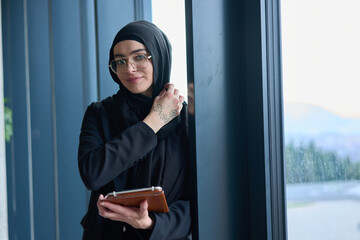 A hijab businesswoman stands by the window in a modern office holding a tablet in her hand...