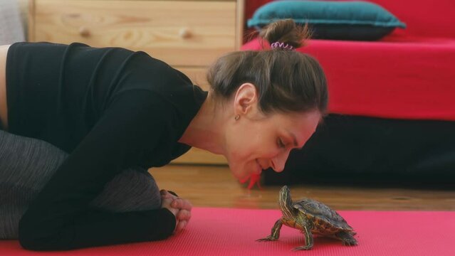 a young cute woman is basking with a red-eared turtle on a yoga mat. Female kisses a pet. The concept of human-animal friendship. High quality 4k footage. Cute funny video