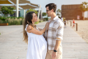 Beautiful young pregnant couple embracing and looking to each other on the beach walkway.