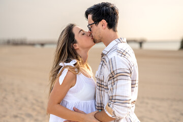 Side view photo of young couple kissing on the beach coastline at sunset.