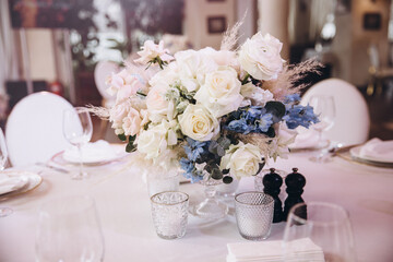 Wedding. Decor. Floristics. The festive table is decorated with a composition of pink and blue flowers with leaves of greenery. There are candles, glasses and plates on the table