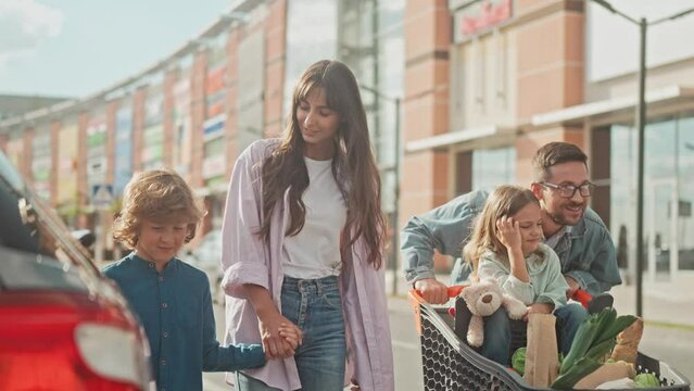 Happy family made groceries at food market. Mom keep son by hand and go to car parking. Dad push daughter in grocery cart. Girl is pointing finger at something to dad. Leisure of children and parents.