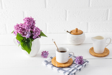 A small white cup of coffee on a wooden white table with a spring bouquet in a jug opposite a white brick wall. Morning coffee.