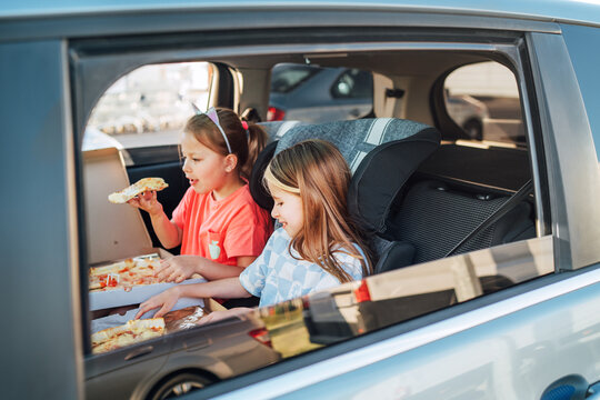 Two little sisters are happy to eat just cooked Italian pizza sitting in child car seats on the car back seat. Happy childhood, fast food eating, or auto journey lunch break concept image.