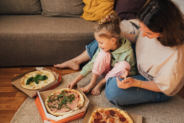 Young mother and her little 5 year old daughter eating pizza together in cozy living room.