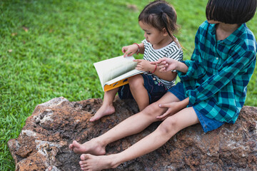 Sister and sister reading a book in the backyard Summer holidays, fairy tales, books and love. Brothers and sisters teach them to sit and read a book on the rocks in their backyard and relax together.