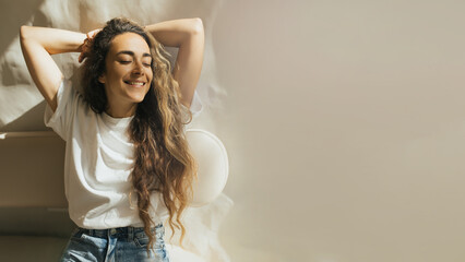 Naklejka premium Young woman with long curly hair in casual clothes sitting near window, smiling.