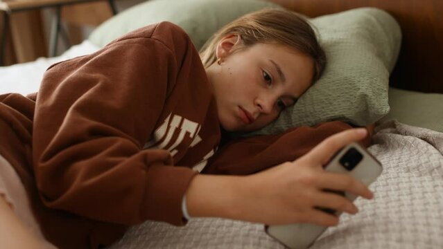 Close up of unhappy teenage girl breathing heavily laying on bed and scrolling through social networks on her smartphone