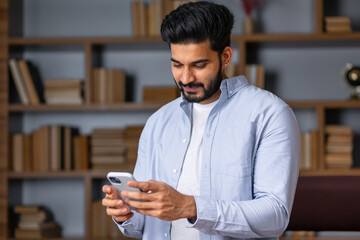 Young indian man holding smartphone tech device using cell phone apps at home. Bearded ethnic guy texting messages looking at smartphone checking social media, ordering online or browsing.