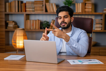 Obraz na płótnie Canvas Millennial arab freelancer man in casual sitting at work desk in front of laptop, gesturing and smiling, having video call with colleagues while working from home, copy space