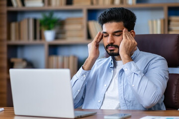 Tired young man, office worker, manager holding his head, feeling pain, dizziness. Sitting at a computer desk in the office.