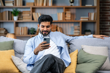 Young indian man holding smartphone tech device using cell phone apps at home. Bearded ethnic guy texting messages looking at smartphone checking social media, ordering online or browsing. - 610647252