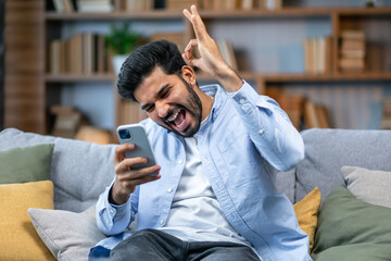 Surprised happy man holding phone looking at cellphone read good news in sms sit on sofa, amazed...