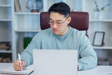 Focused young asian businessman study online watching webinar podcast on laptop listening learning education course conference calling make notes sit at work desk, e-learning concept