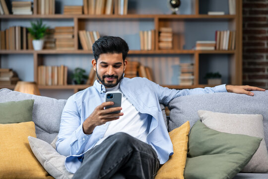 Young indian man holding smartphone tech device using cell phone apps at home. Bearded ethnic guy texting messages looking at smartphone checking social media, ordering online or browsing.