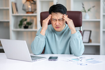 Tired young asian worker at work. He has a headache, keeps his hands on his head, sits at a desk in...