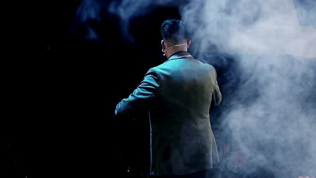 Close-up of magician presenter speaking on stage with microphone and smoke behind him