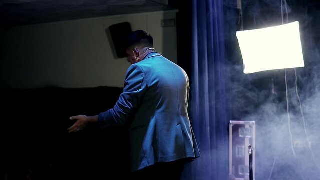 Magician presenter speaking in slow motion on small stage with smoke. View from the audience