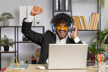 Happy relaxed overjoyed indian businessman working on laptop computer at home office wearing headphones listening favorite energetic disco music and dancing. Freelancer man relaxing, taking a break