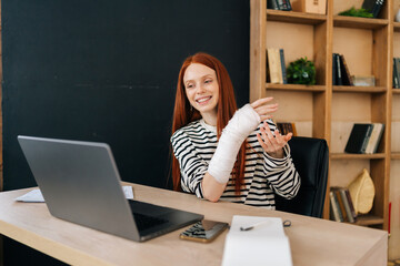 Fototapeta Cheerful young woman with broken right hand wrapped in white gypsum bandage having online video call, smiling looking to screen, working remotely on laptop computer sitting at desk in home office room obraz