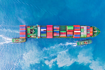 Aerial top view of container ship cruising in Mediterranean port with deep blue sea assisted by tug boat.