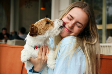 Wire haired jack russell terrier licking a cheek of his joyful woman outside of the coffee shop. Young woman and her dog playing outdoors. Copy space, background. - obrazy, fototapety, plakaty