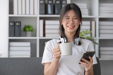 Fototapeta premium Attractive and happy young Asian female using her smartphone to scroll through social media while relaxing on her comfortable sofa in her living room.