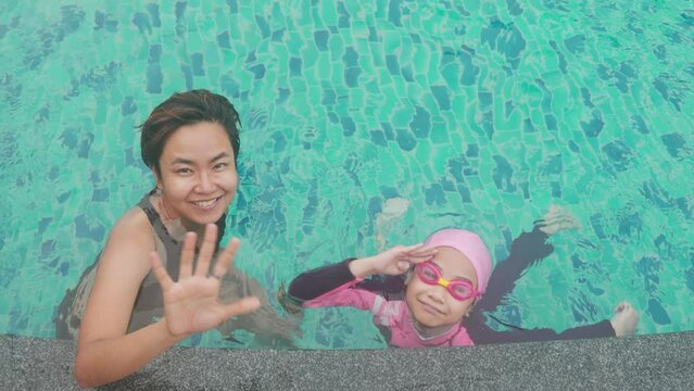 Beautiful asian mother and daughter who are swimming in swimming pool with smile, happiness and bye-bye hands shows concept of relaxation and enjoying their holiday vacation in summer time.