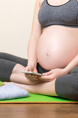 Fototapeta Pregnant female holding smartphone while sitting on exercise yoga mat at home at coronavirus time obraz