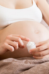 a pregnant girl sits at home on the bed and smears smile an anti-stretch mark cream on her stomach. Pregnancy, motherhood, preparation and expectation concept