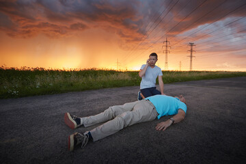 Young man calling emergency services for unconscious middle aged man. Dramatic resuscitation on rural road under storm clouds. Themes rescue, help and hope.. - Powered by Adobe