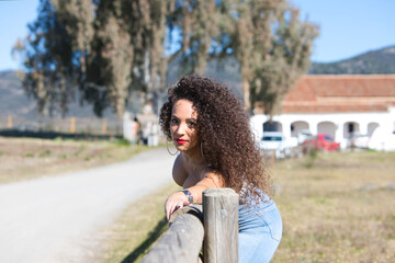 Curly-haired brunette woman has her arm stretched over the wooden fence of the path leading to the meadow as she looks at the camera. Dressed in jeans and a white top, she enjoys the sunny day.