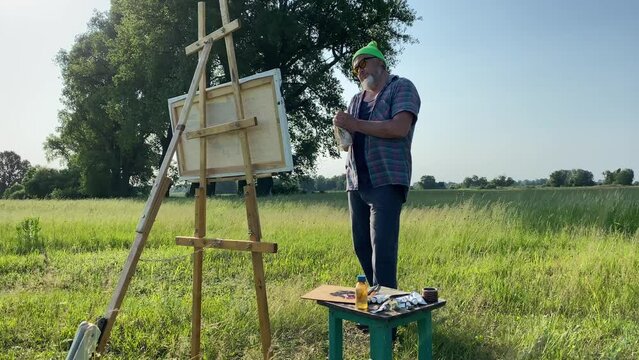 Dolly shot jolly artisan enjoying result of his inspiration, contemplates oil painting while cleaning hands with rag, standing at meadow in sunny beams of morning time. Enjoyable painter finished work