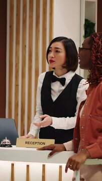 Vertical Video Helpful asian receptionist presenting holiday package options to african american couple in luxury hotel. Husband and wife on honeymoon trip requesting special room amenities in resort