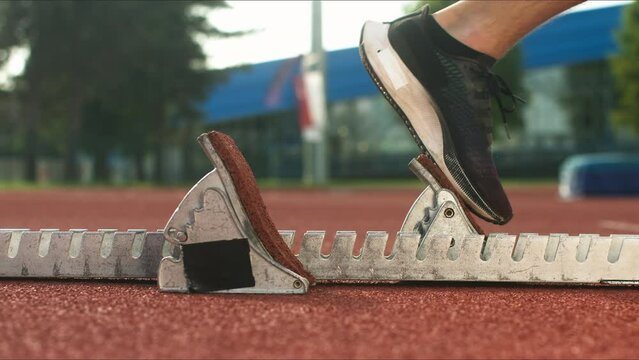Male Caucasian sprinter legs in black sneakers attending a crouching position in the starting block, sideways close up shot.