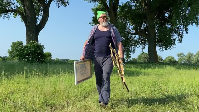 Cool middle-aged artist looks for the best scenic view, prepares for creative process in solitude at nature in sunny morning. Stylish man walks at field, holds easel and canvas. Full length front view