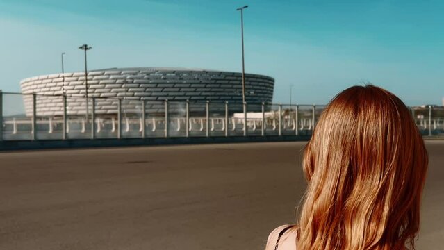 A girl walks down the street and looks at a white stadium against a blue sky, filming with her back. High quality 4k footage