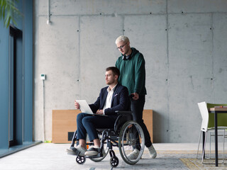 A businessman in a wheelchair in a fashionable office using a laptop while behind him is his business colleague who gives him support