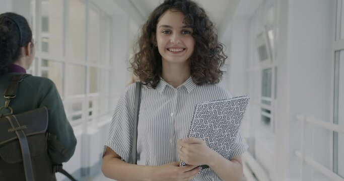 Portrait of cheerful girl standing in lobby of modern university holding textbooks smiling while students walking by. Youth and education concept.