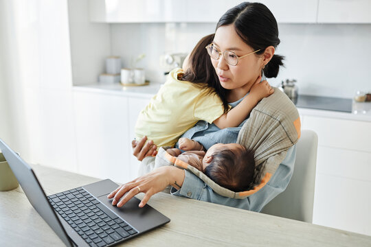 Asian Young Mother With Two Kids Working Online On Laptop While Sitting In The Kitchen