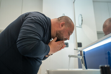 Fototapeta Fat, bald, bearded middle-aged man washes face in bathroom home int sink. Overweight and sweat. Daily routine, hygiene procedure, morning care and body cleanliness concept obraz