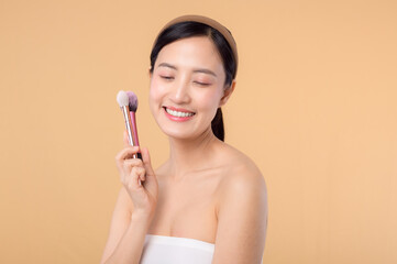 Portrait happy young asian woman with natural make up face holding cosmetic skin powder blusher isolated on beige background. Female apply skincare brush treatment. beauty product cosmetology concept.