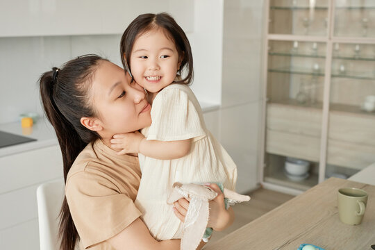 Asian young mom embracing her little daughter while they sitting in the kitchen